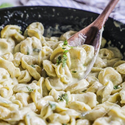 a wooden spoon scooping tortellini with pesto cream sauce and fresh basil out of a large, black skillet.