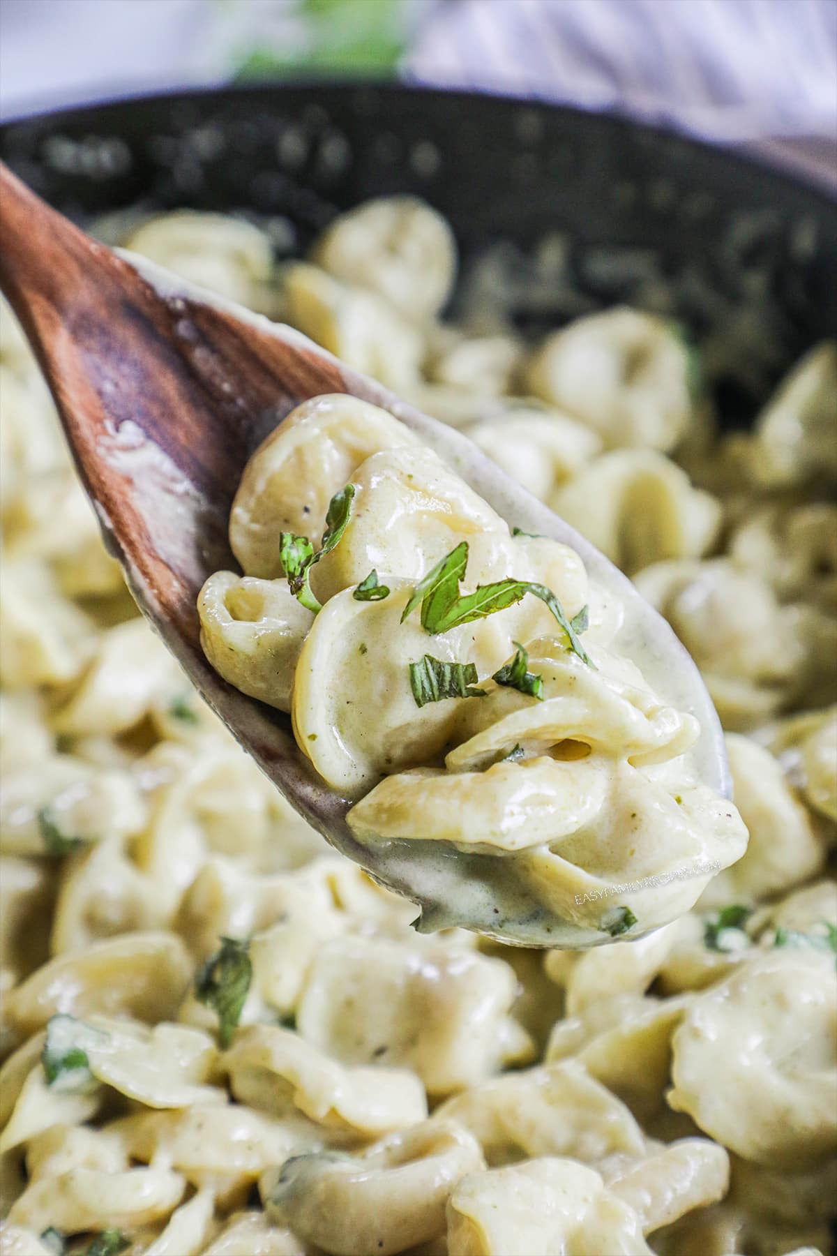 a wooden spoon scooping tortellini with pesto cream sauce and fresh basil out of a large, black skillet.