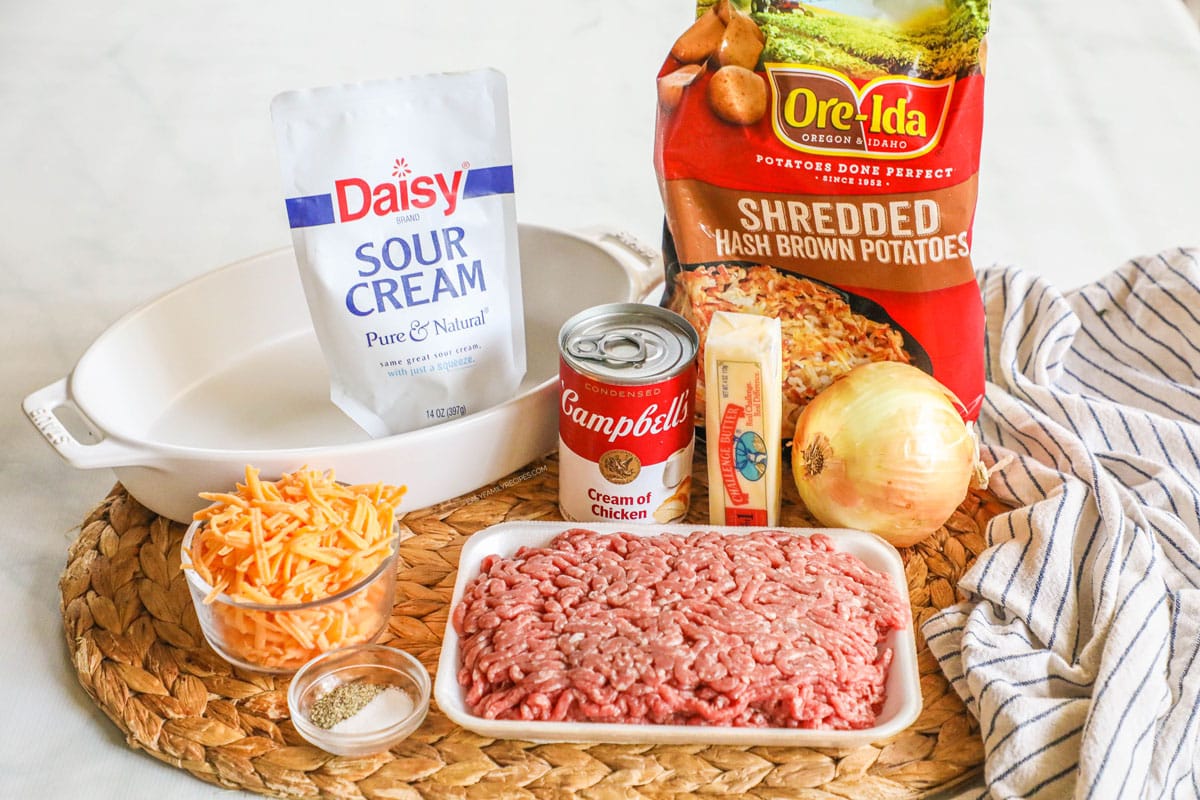 Ingredients for ground beef hashbrown casserole, including sour cream, hashbrowns, onion, beef, butter, cream of chicken soup, cheese, salt and pepper