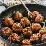 Spoon lifting two honey sriracha meatballs from cast iron skillet