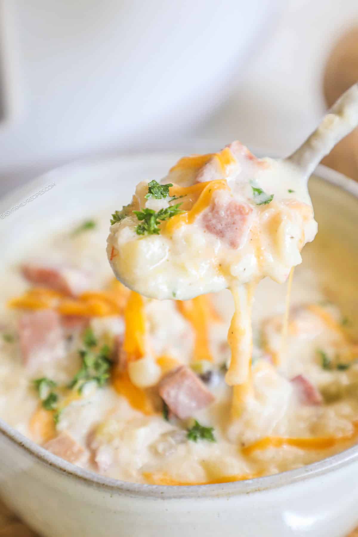 A spoon scooping up a bite of creamy potato soup with ham that's topped with cheddar cheese.
