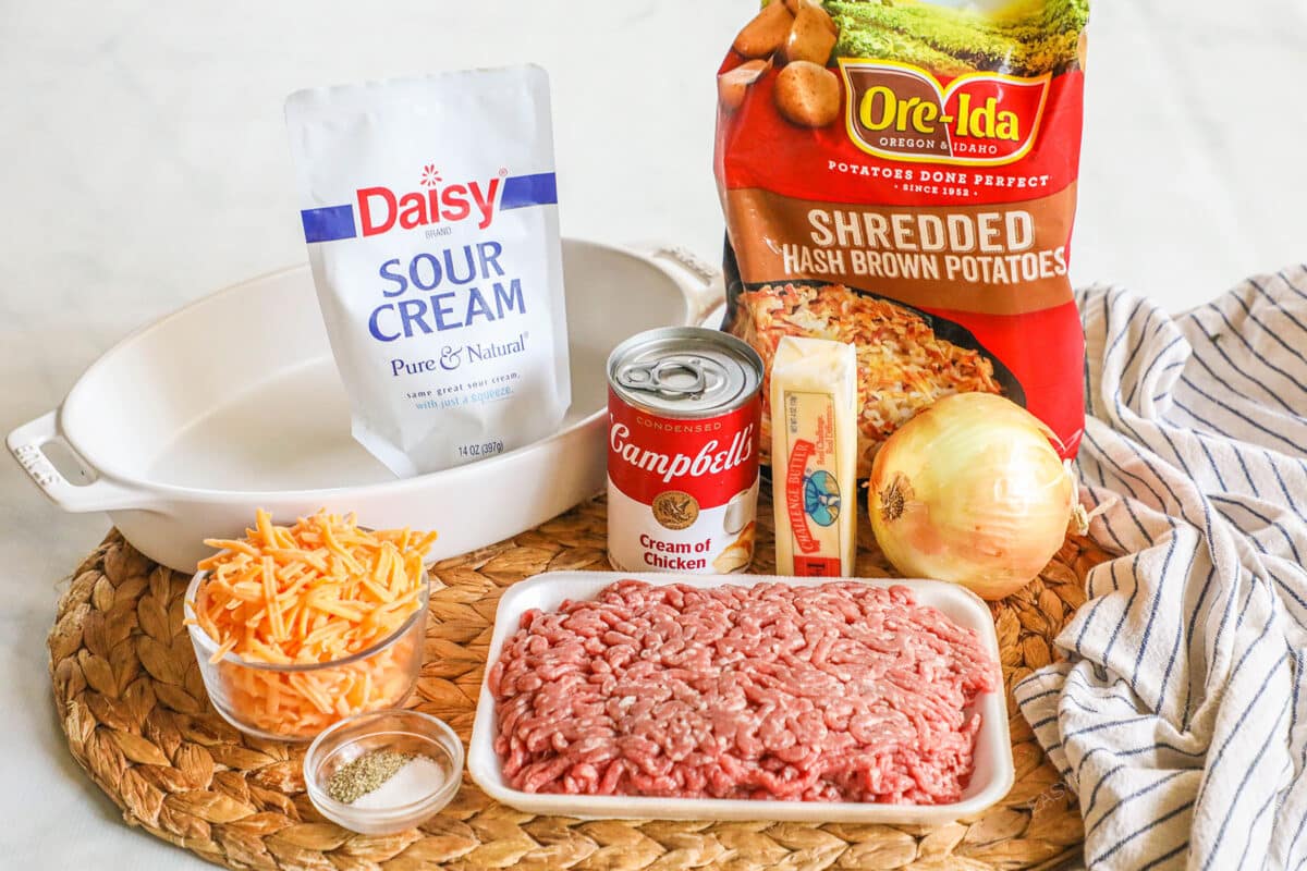 Ingredients for ground beef hashbrown casserole, including sour cream, hashbrowns, onion, beef, butter, cream of chicken soup, cheese, salt and pepper