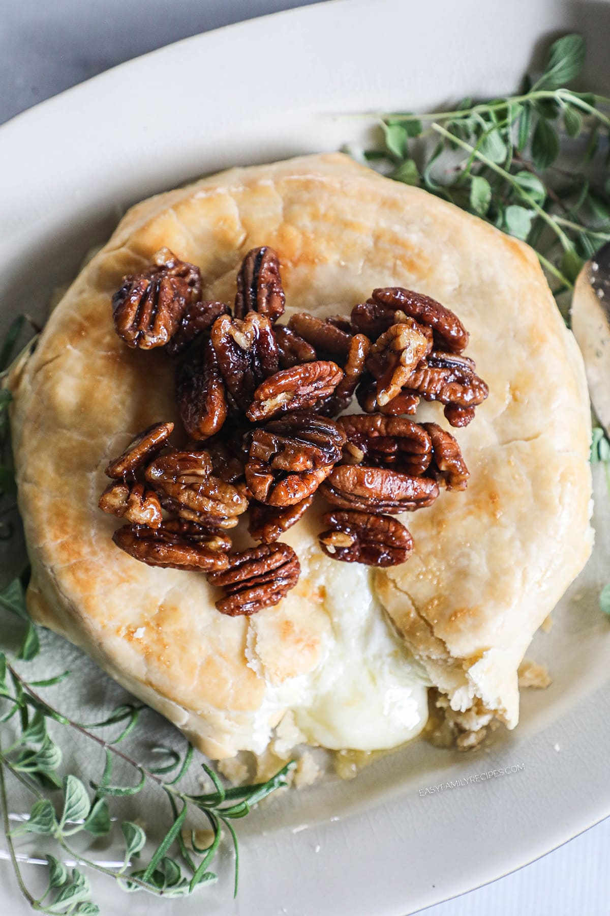 overhead of a wheel of baked brie in a golden pie crust topped with fresh herbs and pecans.