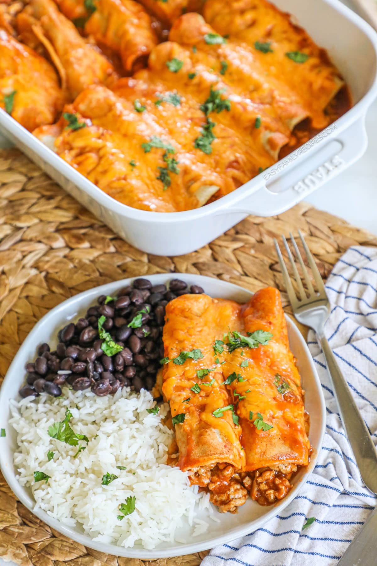 Turkey enchiladas on a plate with black beans and rice with the casserole dish of enchiladas nearby.