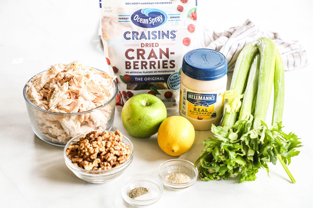 Ingredients for recipe before prepping: a bowl of shredded chicken, walnuts, an apple, a lemon, bunch of celery, mayo, dried cranberries, and spices.