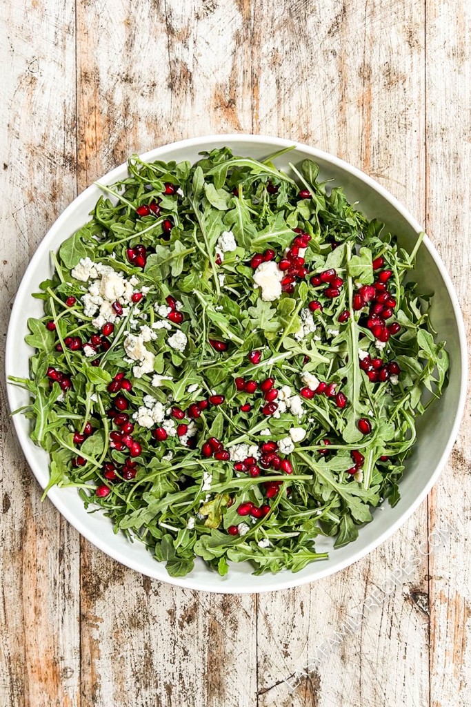 above image of arugula topped with cheese and pomegranate seeds.