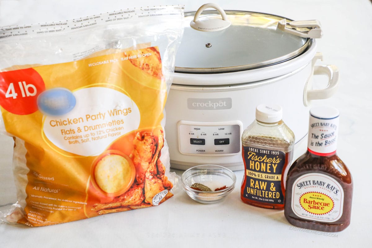 Ingredients for Crockpot honey BBQ chicken wings including frozen chicken party wings, spices, honey, and barbecue sauce. Equipment is a white Crockpot with lid. 