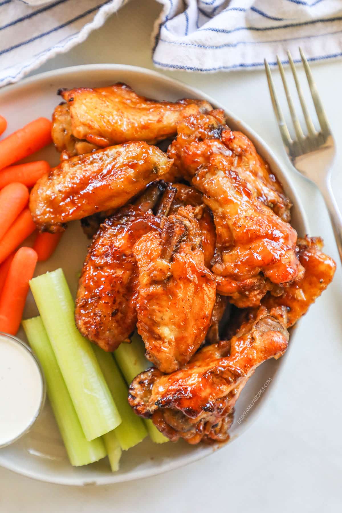 Honey BBQ chicken wings are served on a white plate alongside baby carrots, celery, and ranch dip. A fork and blue and white striped napkin are adjacent. 