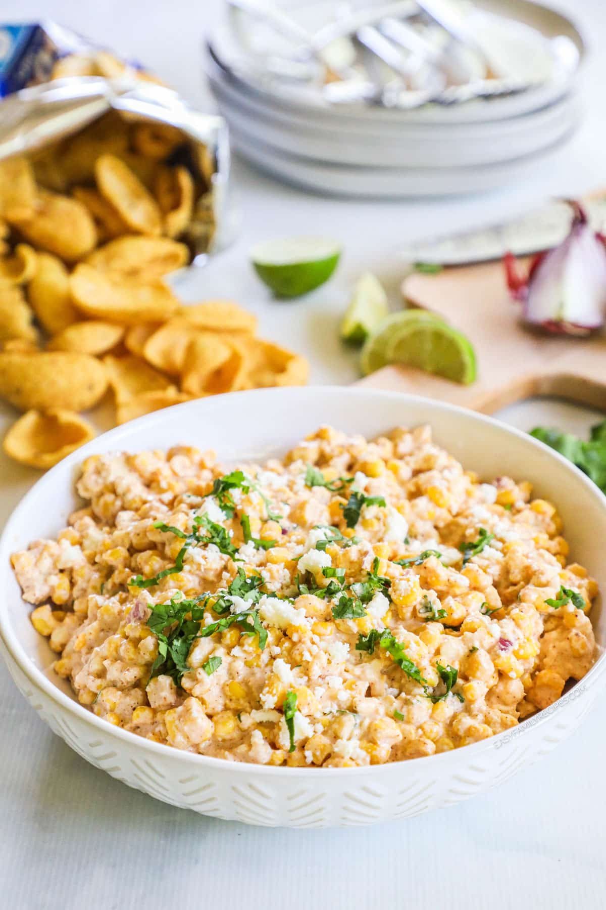 Mexican street corn dip in a serving bowl garnished with cheese and cilantro. Open bag of frito chips and lime wedges nearby.