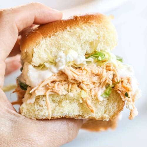 A hand holding a Hawaiian roll sandwich with shredded buffalo chicken, lettuce, blue cheese, and ranch dressing.