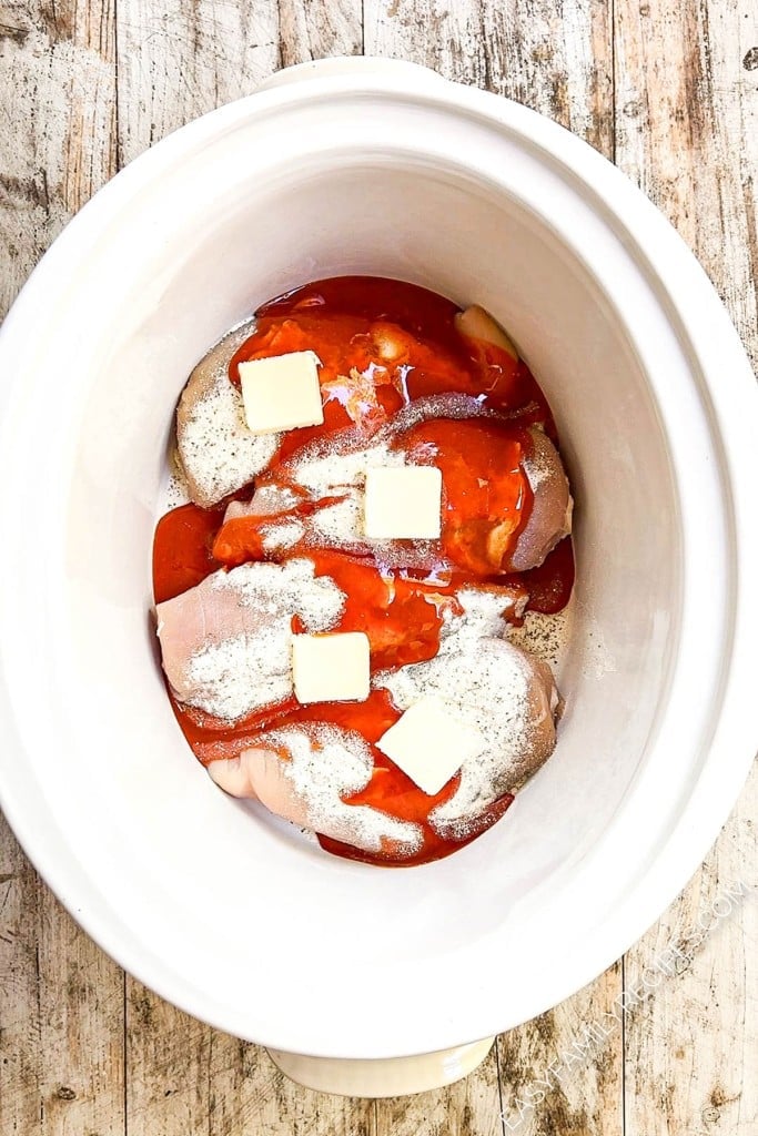A Crockpot with chicken breasts, ranch seasoning, buffalo sauce, and butter inside.