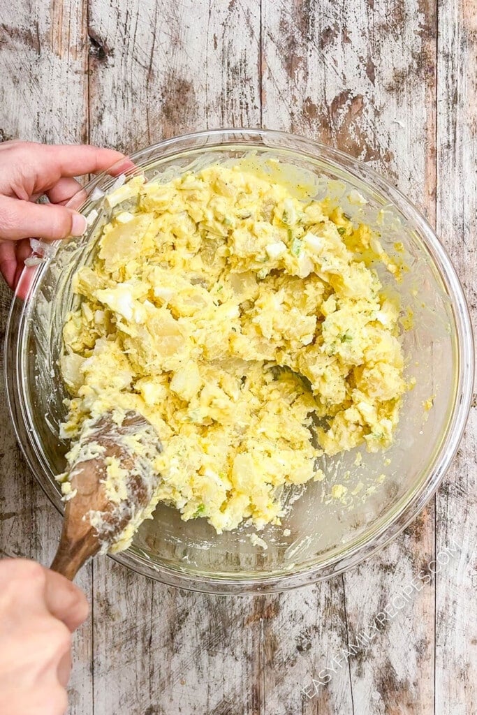Hand stirring together potato salad ingredients in a bowl to fully combine.