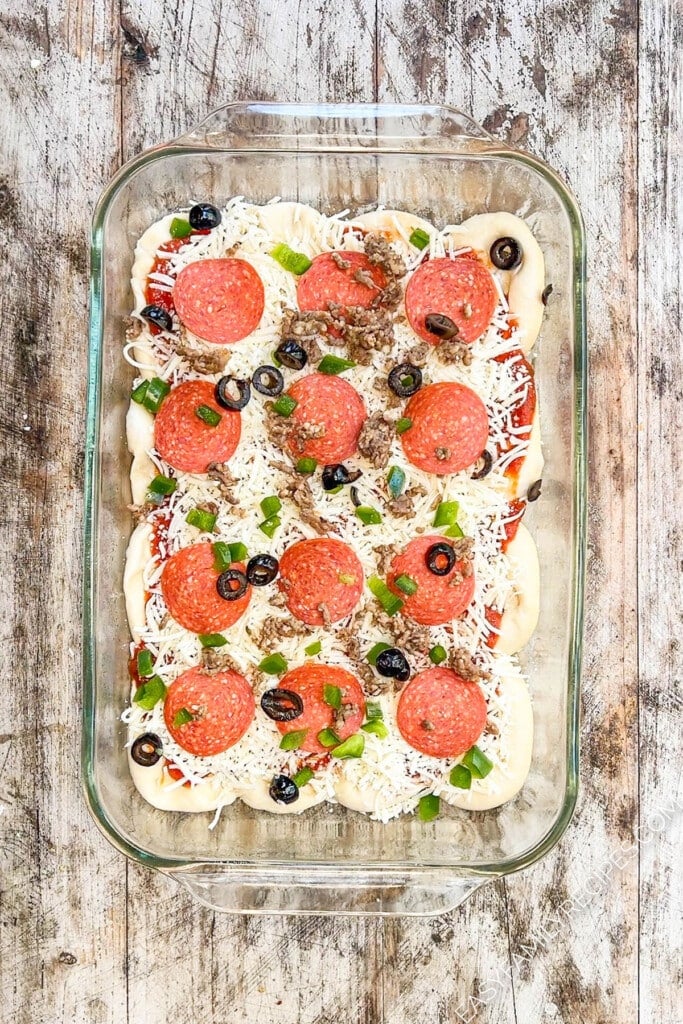 an unbaked pizza with pepperoni, green peppers, olives, and cheese in a rectangle clear baking dish.