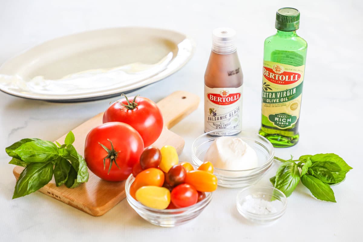 Image of ingredients to make burrata caprese salad - yomatoes, burrato, olive oil basil, aslt and balsamic glaze