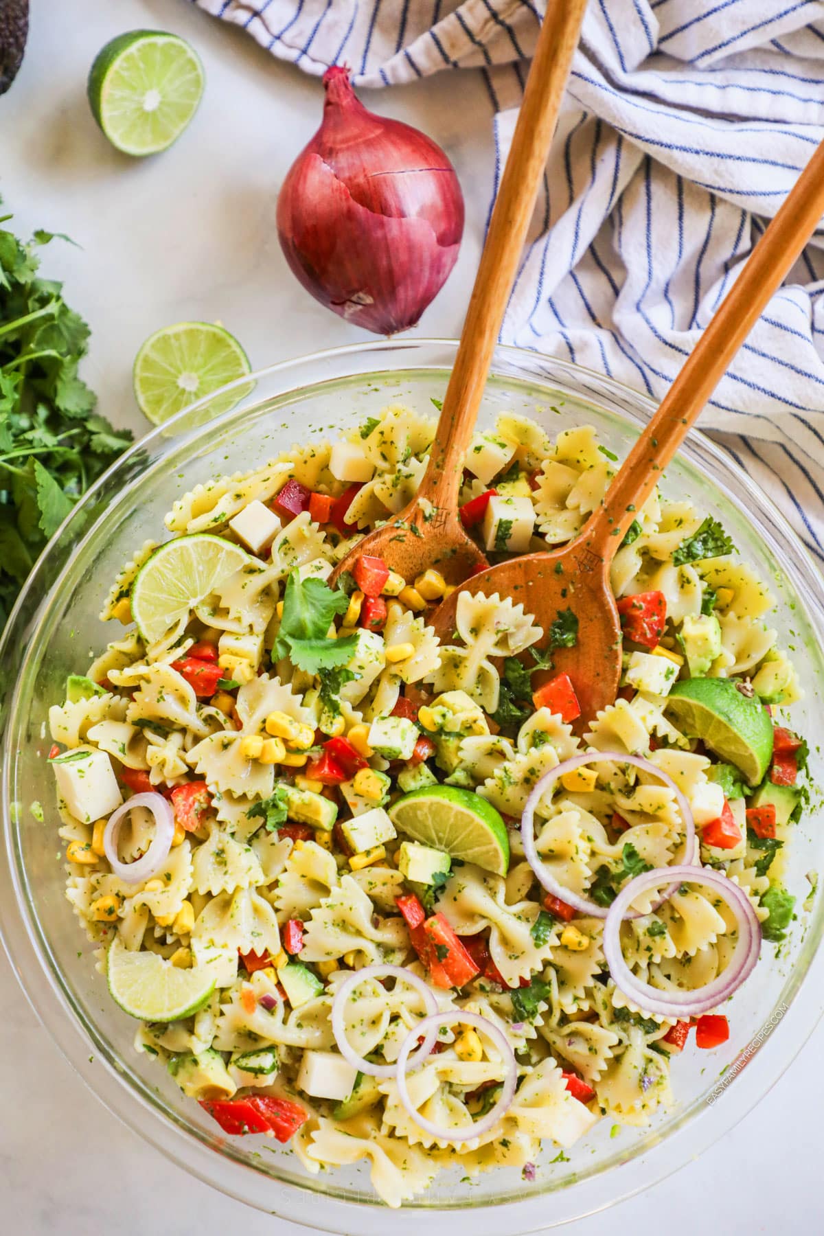 Two wooden spoons in a large bowl of cilantro lime pasta salad made with farfalle, corn, peppers, and cheese.