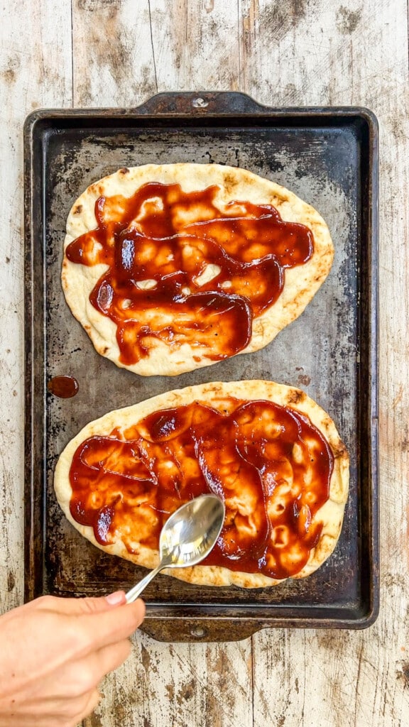 two flatbreads on a baking sheet spread with bbq sauce.