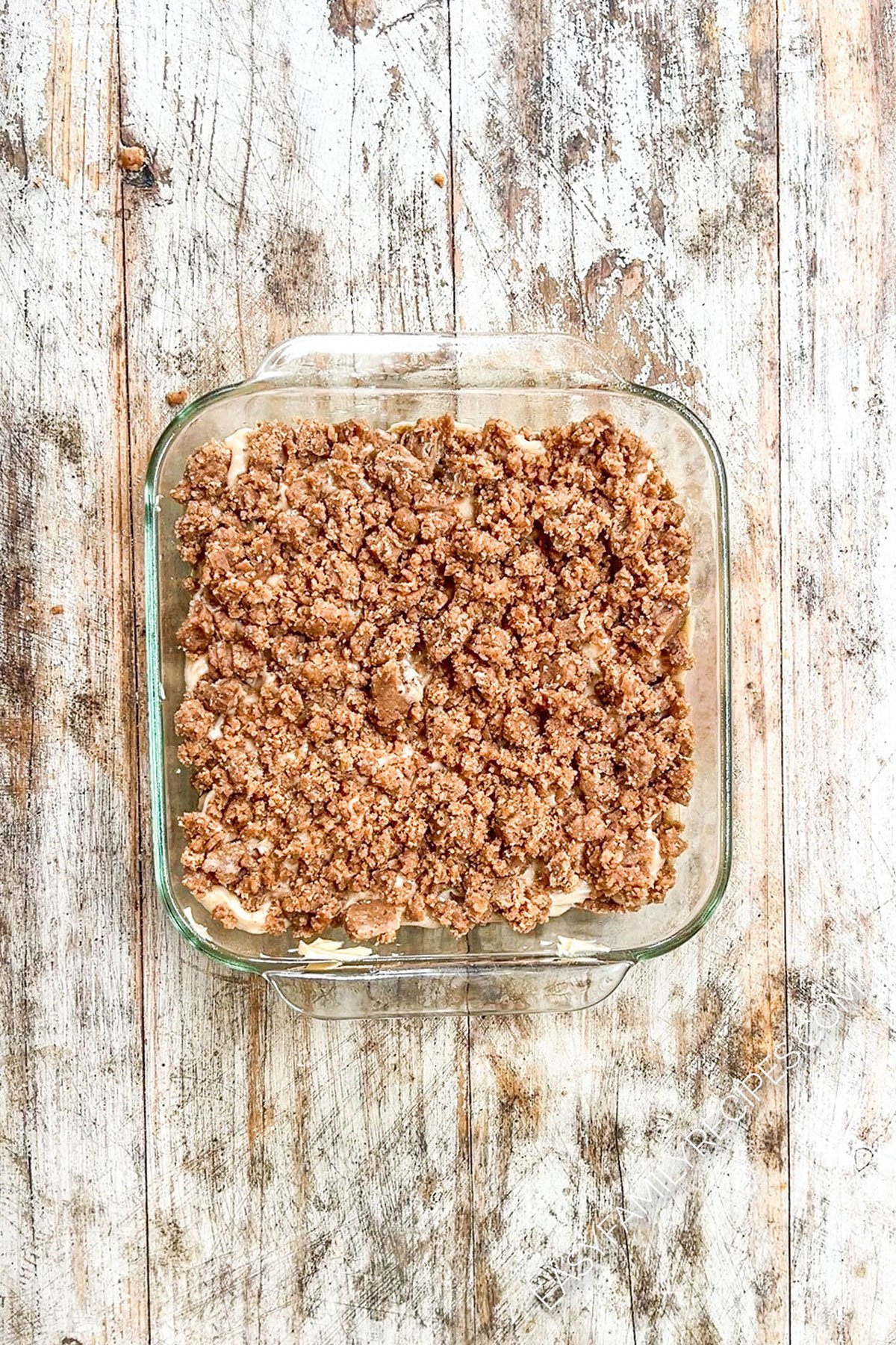 A glass baking dish half full of cinnamon crumble coffee cake batter, with the cinnamon crumble layer being added.