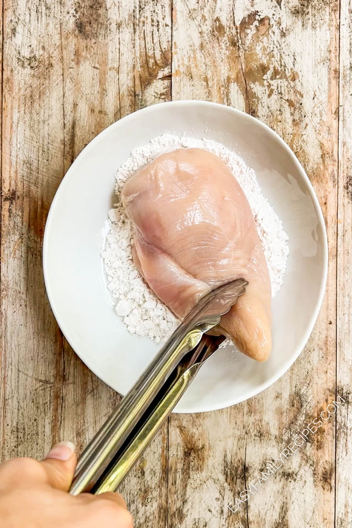 Tongs dipping a chicken breast into a bowl of flour.