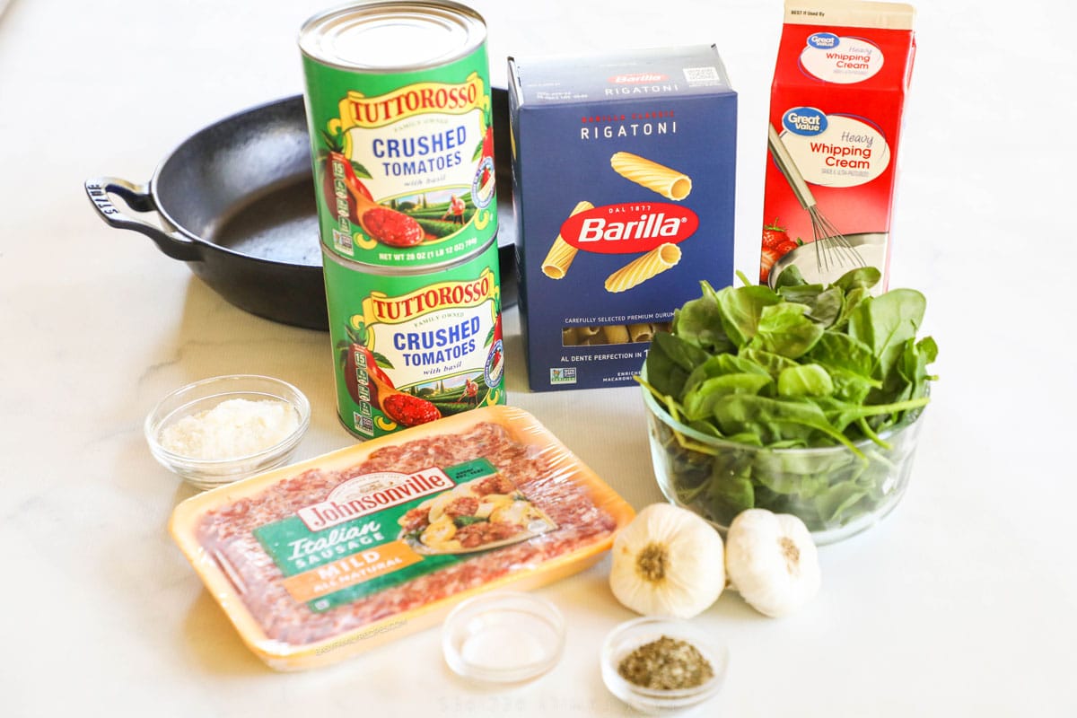 Ingredients to make creamy Italian sausage pasta including sausage, spinach, canned tomatoes, cream, and dried pasta