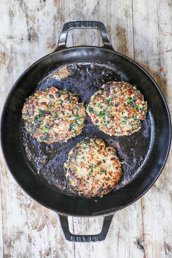 Tuscan turkey burger patties cooking on a cast-iron pan with a distressed wood background.