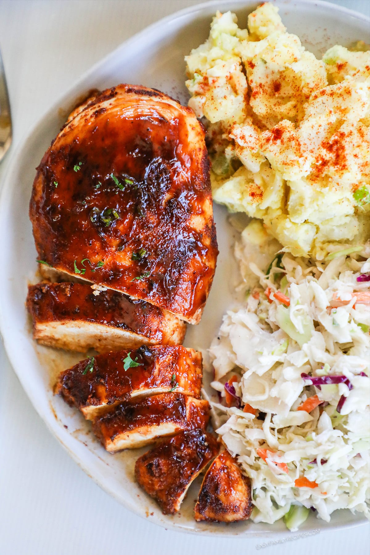 a partially sliced oven baked BBQ chicken breast on a plate with coleslaw and potato salad from above.