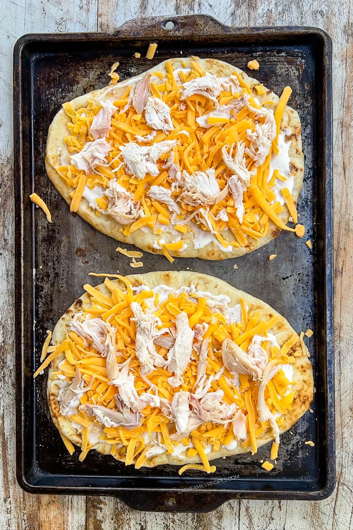 Two buffalo chicken flatbreads are on a baking sheet, with cheese and chicken.