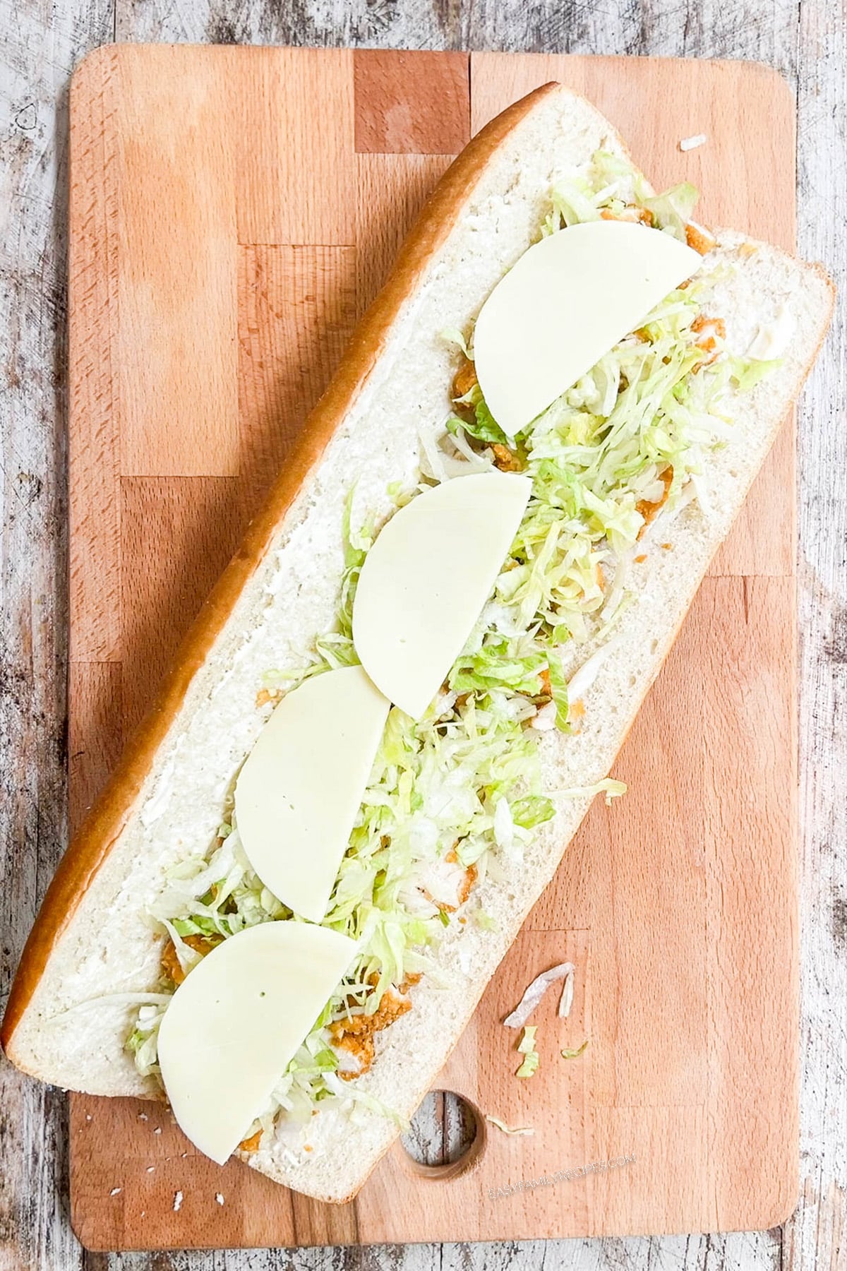 Assembling a chicken tender sub on a wooden cutting board: Adding the lettuce and cheese.
