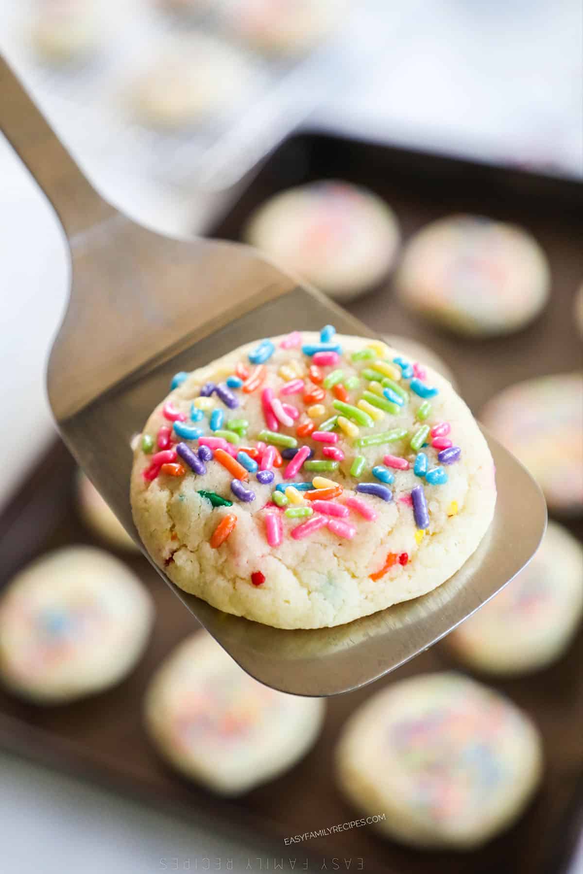a birthday cake cookie being held on a spatula over a baking sheet filled with cookies.