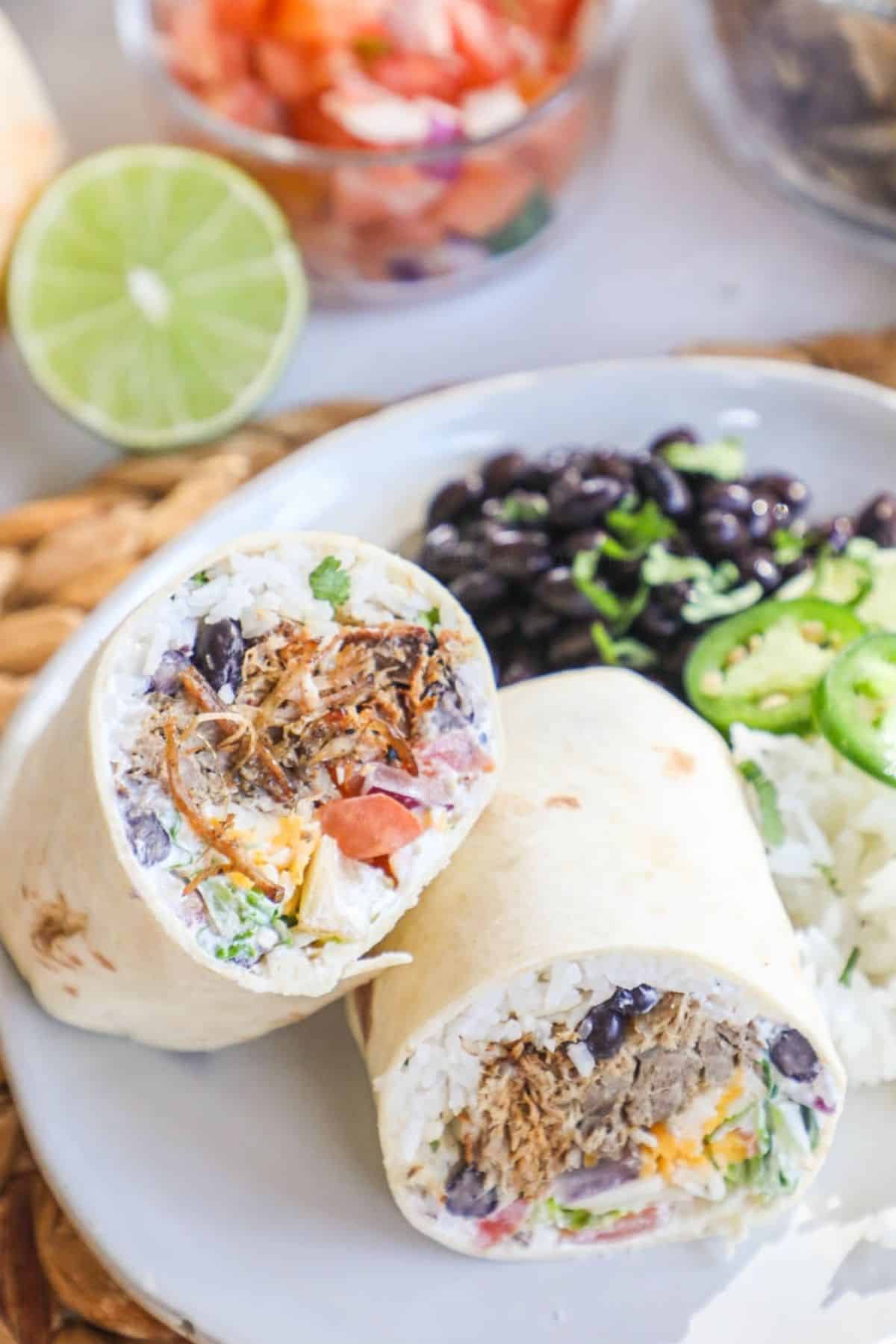 2 carnitas burrito halves on a plate with white rice and black beans