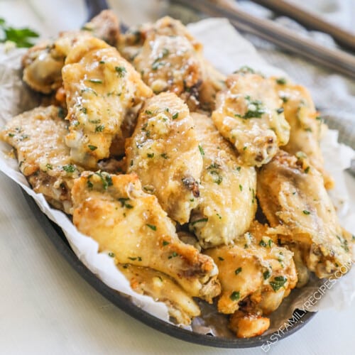 A parchment paper lined tray piled with slow cooker garlic Parmesan wings, garnished with extra cheese and herbs. Tongs are in the background.