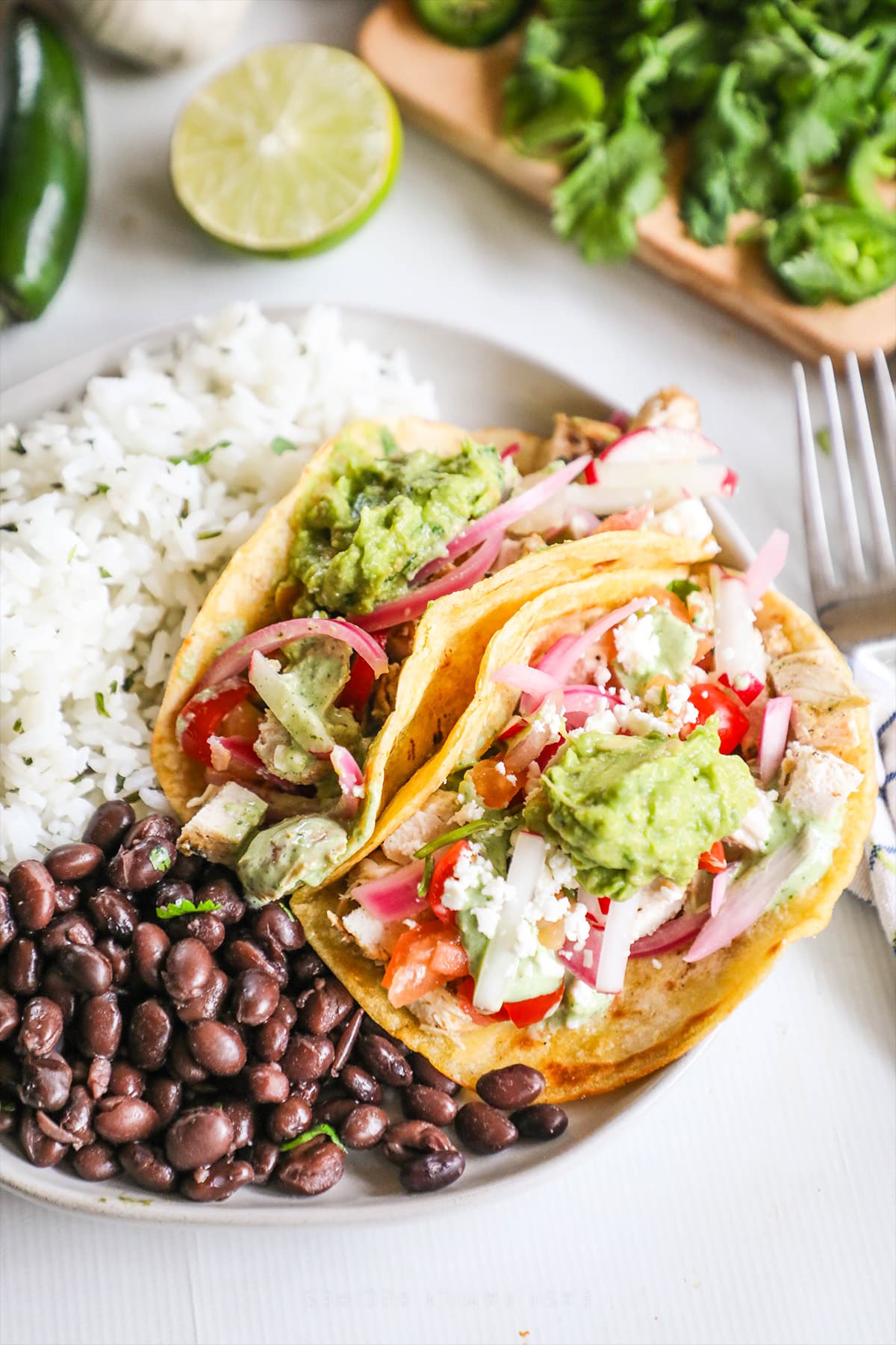 A white plate with two Baja Chicken Tacos along with beans and rice for a side. The plate sits on a white background with a fork, lettuce, lime, and jalapeño adjacent.