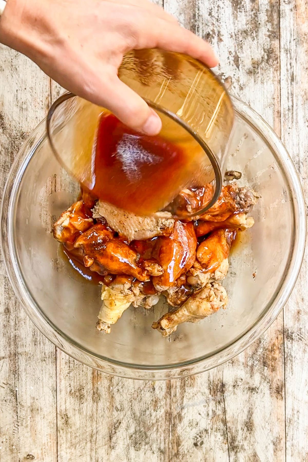 A clear glass bowl full of cooked wings, with Asian Zing sauce being poured over the top.