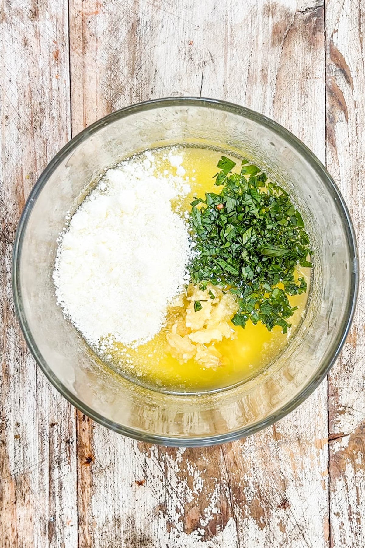 Ingredients for slow cooker garlic Parmesan wings are added to a clear glass bowl with a distressed white wood background.