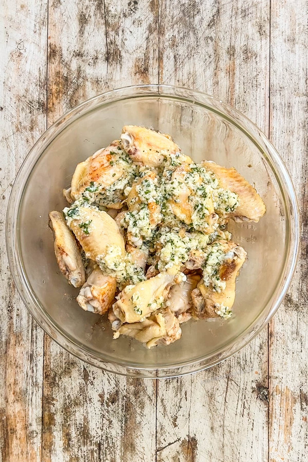 Slow cooker garlic Parmesan wings being tossed with ingredients in a clear glass bowl with a distressed wood background.