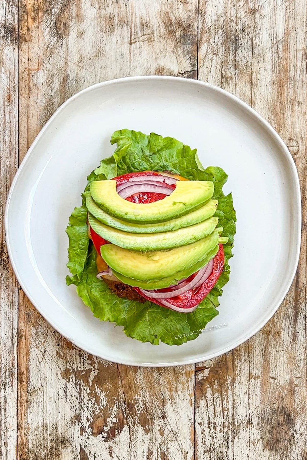 Cooked burger on a bottom bun with lettuce, onion, tomato and sliced avocado.