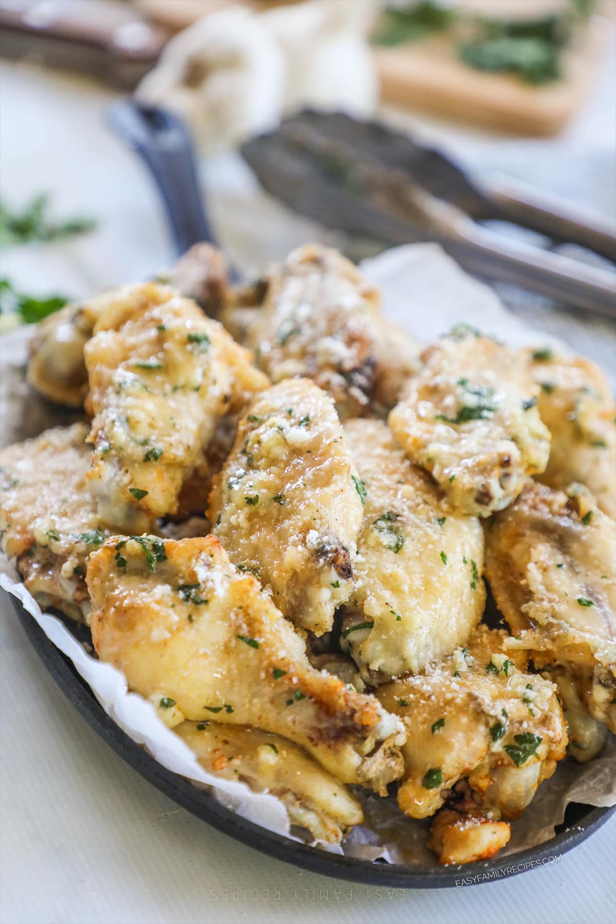 A parchment paper lined tray piled with slow cooker garlic Parmesan wings, garnished with extra cheese and herbs. Tongs are in the background.