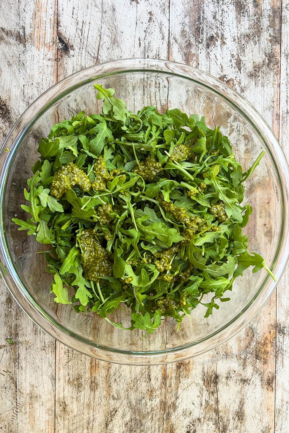 A clear glass bowl with arugula being tossed with pesto for arugula burrata salad.