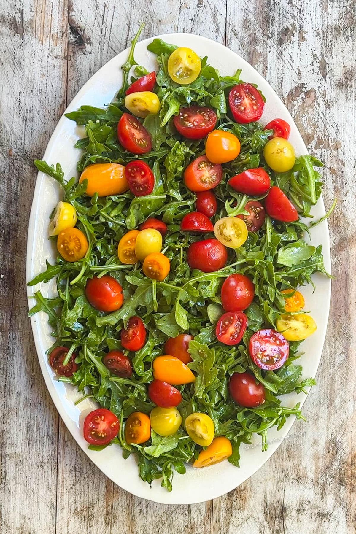 Top view of arugula burrata salad in process, with the arugula tossed in pesto and topped with cherry tomatoes, on a white plate with a distressed wood background.