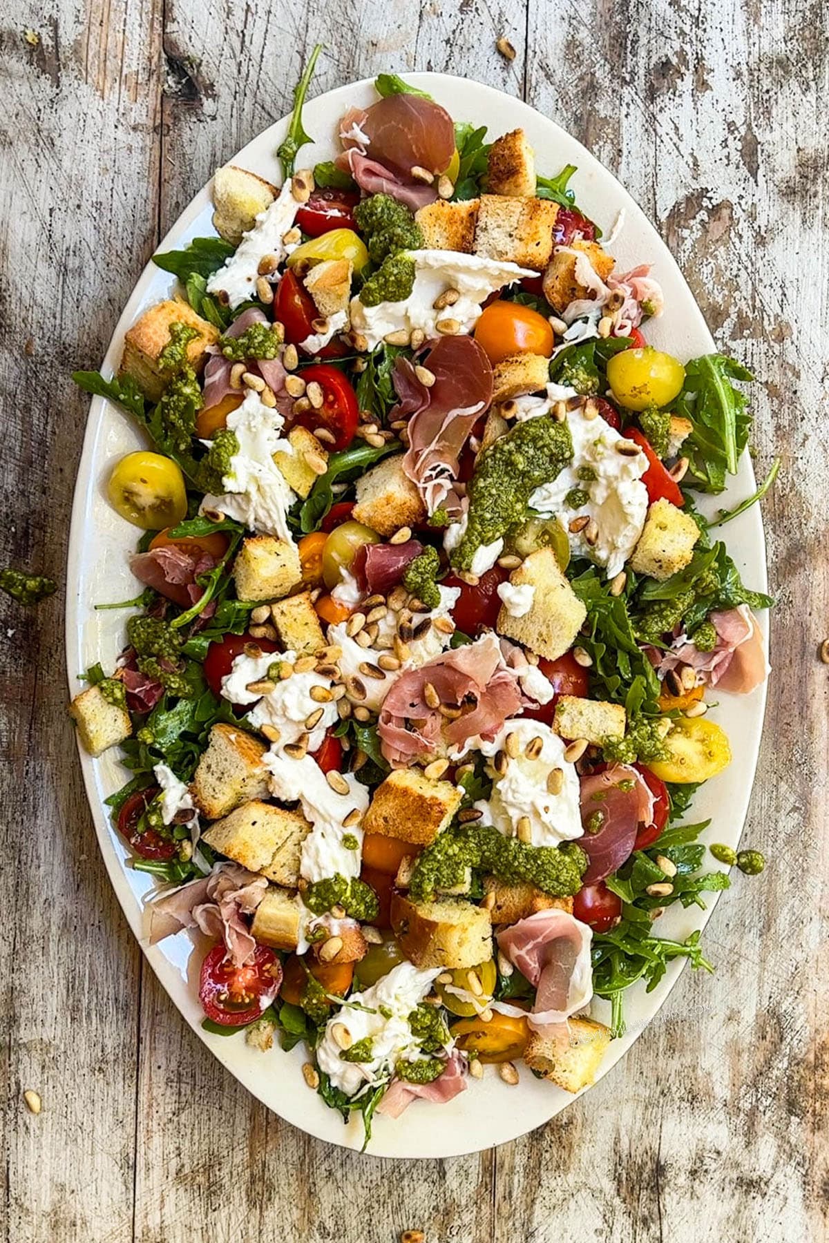 Top view of a white plate full of arugula burrata salad, with arugula, cherry tomatoes, pesto, croutons, prosciutto, and burrata.