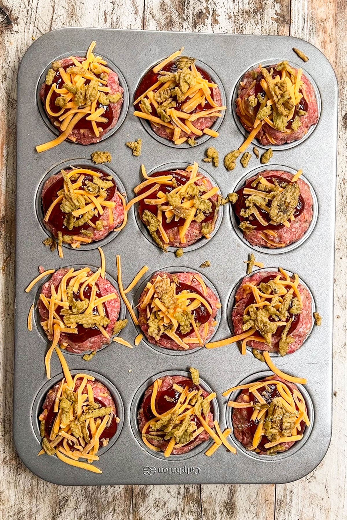 Meat mixture divided into a 12 tin muffin pan with bbq sauce and cheese on top, before baking.