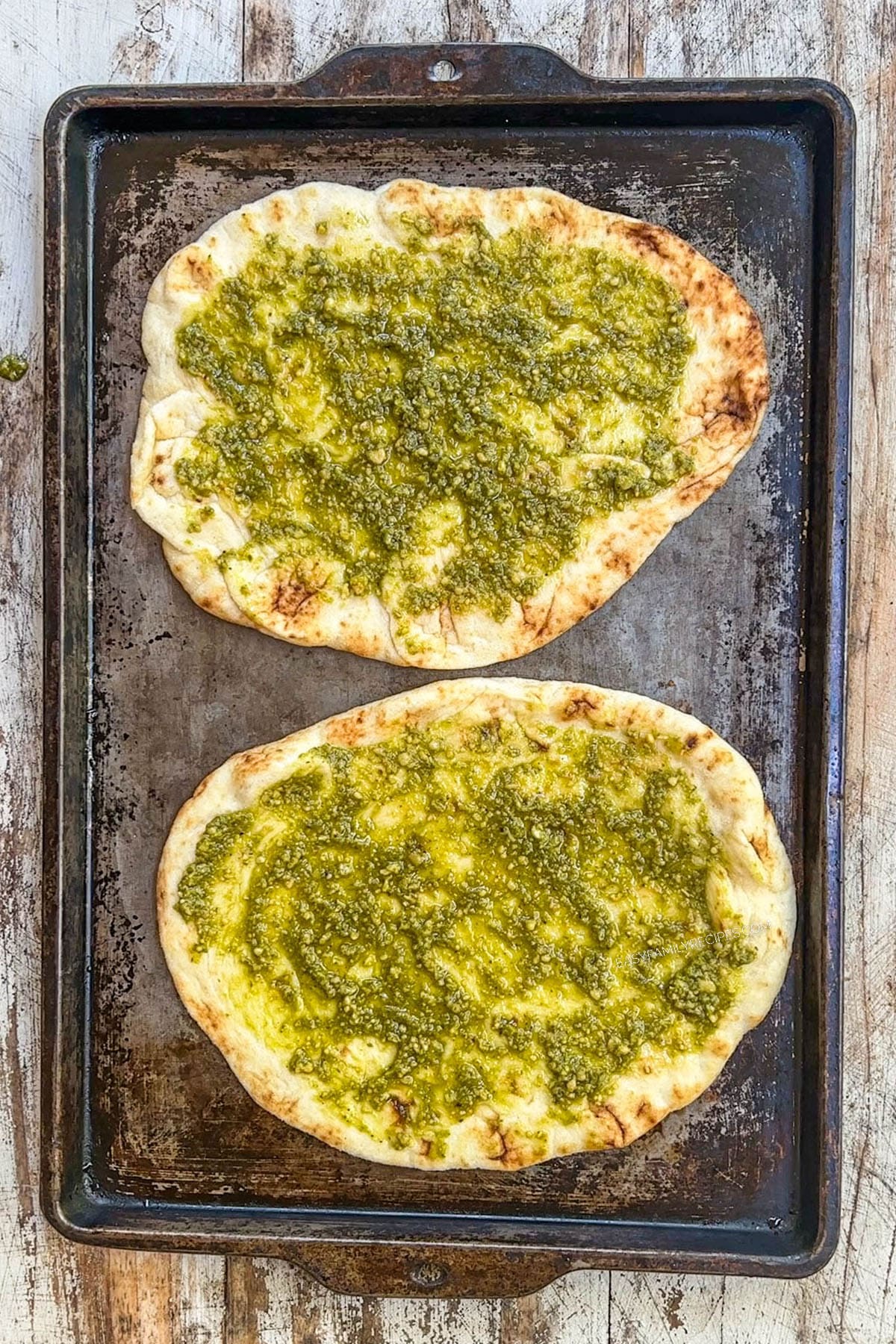 2 flatbreads on a sheet pan with pesto sauce spread over them.