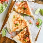 A top view of Chicken Bacon Ranch Quesadillas on a piece of parchment paper with lime wedges and veggies scattered around. The text reads, "Chicken Bacon Ranch Quesadilla"