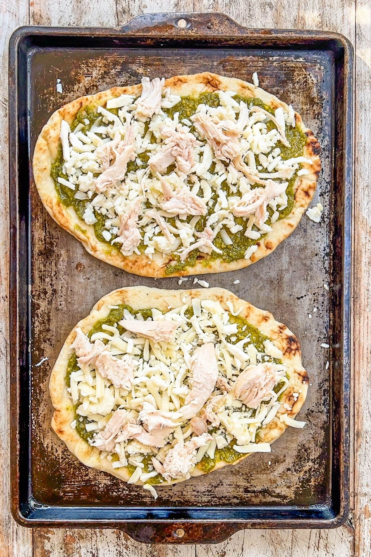 Pesto, cheese, and chicken are added to Mediterranean Flatbread pizzas on a baking sheet.