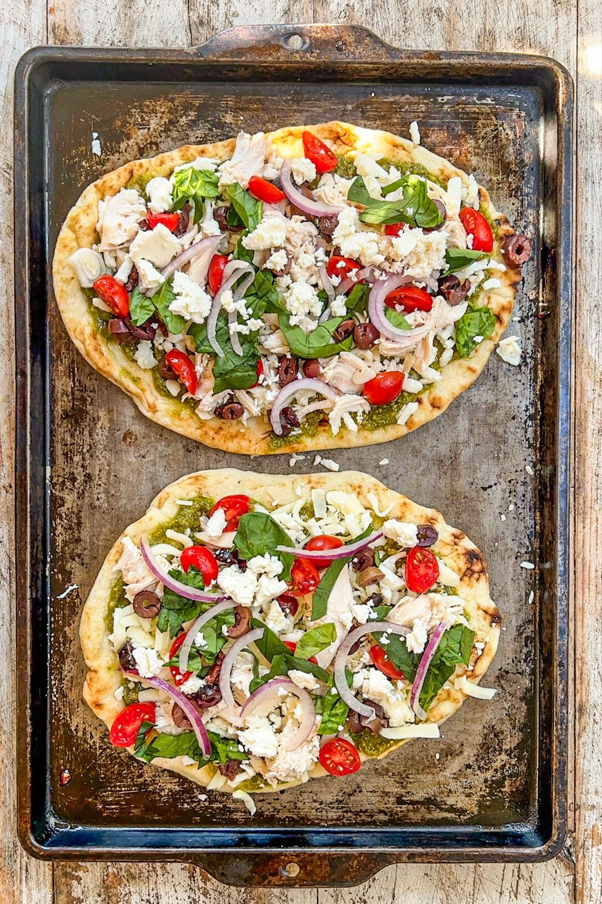 A top view of two Mediterranean Chicken Flatbread pizzas being assembled on a baking sheet.