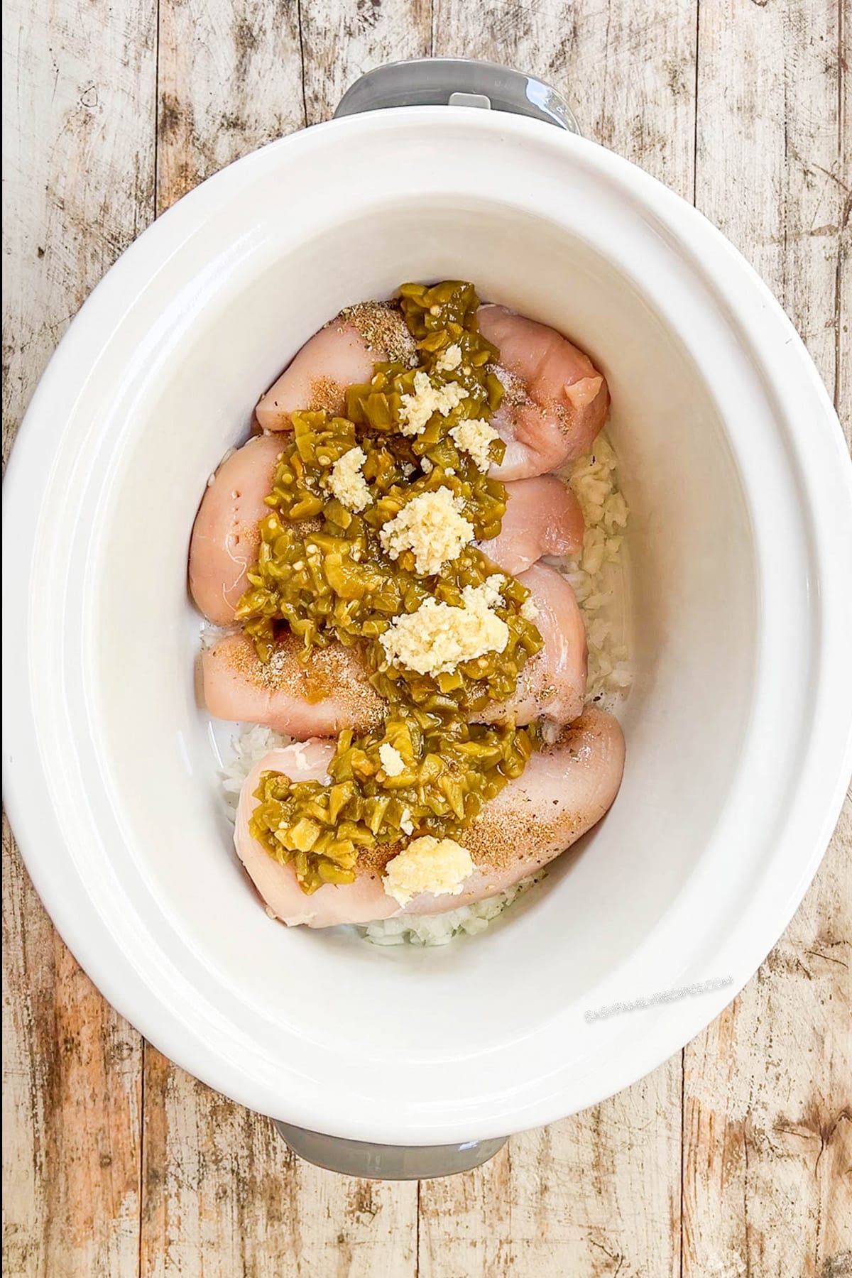 A top view of a white Crock Pot with ingredients for Salsa Verde Chicken Soup being added; step 1 of making the soup.