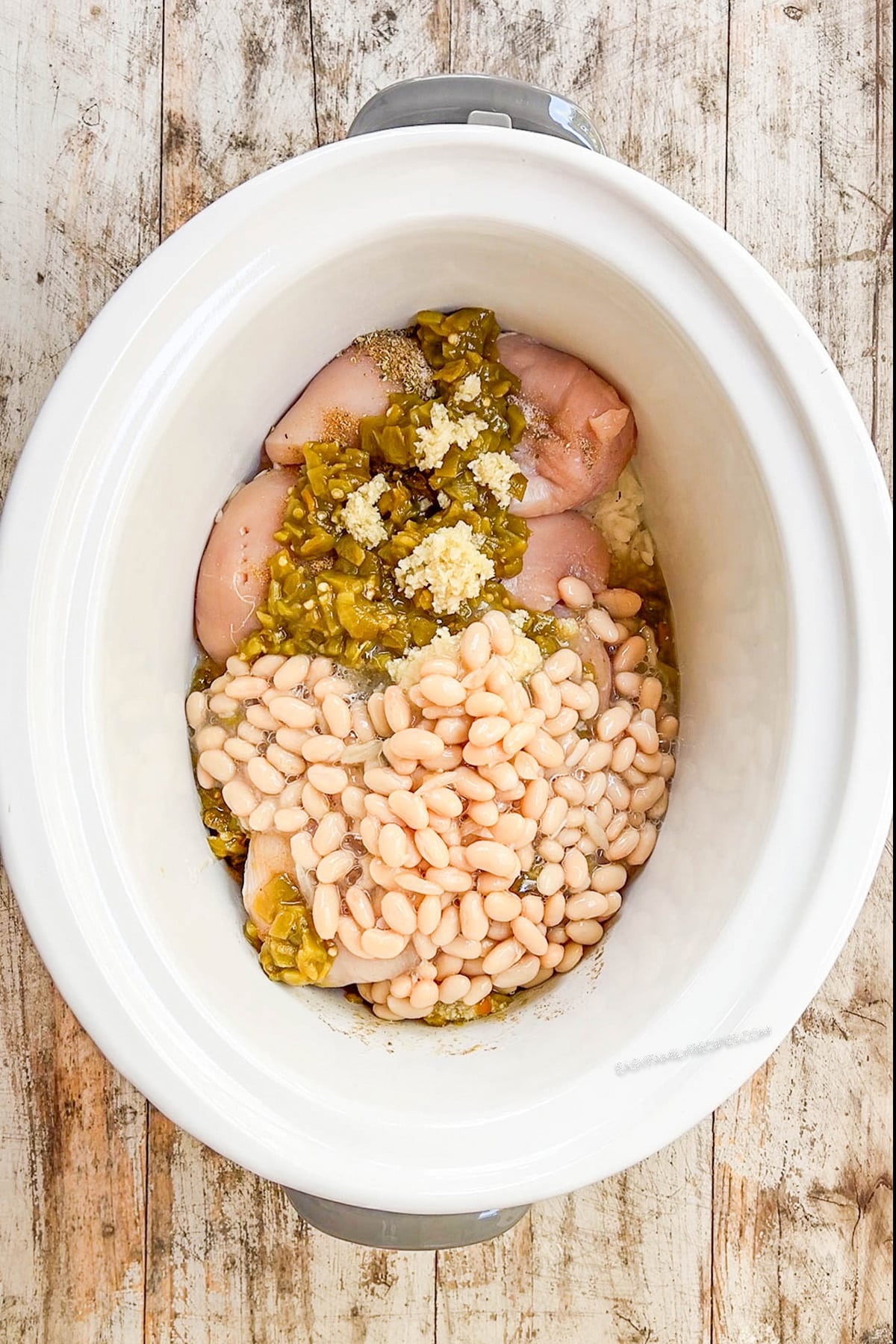 A top view of a white Crock Pot with beans and other ingredients for Salsa Verde Chicken Soup being added; step 2 of making the soup.