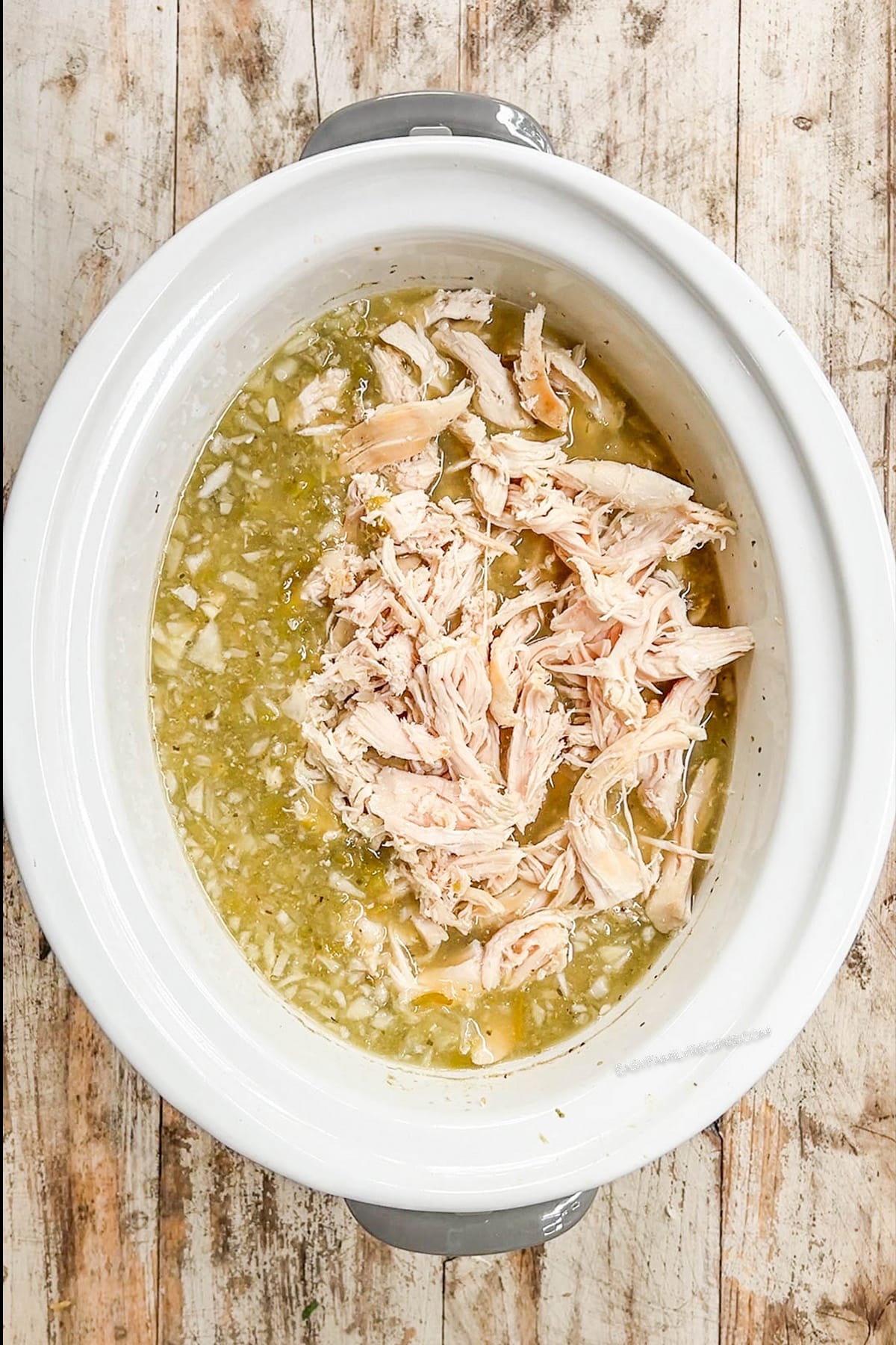 A top view of a white Crock Pot with the cooked chicken for Salsa Verde Soup. The chicken is shredded and the soup is just about ready to serve. Step 5 of the recipe.