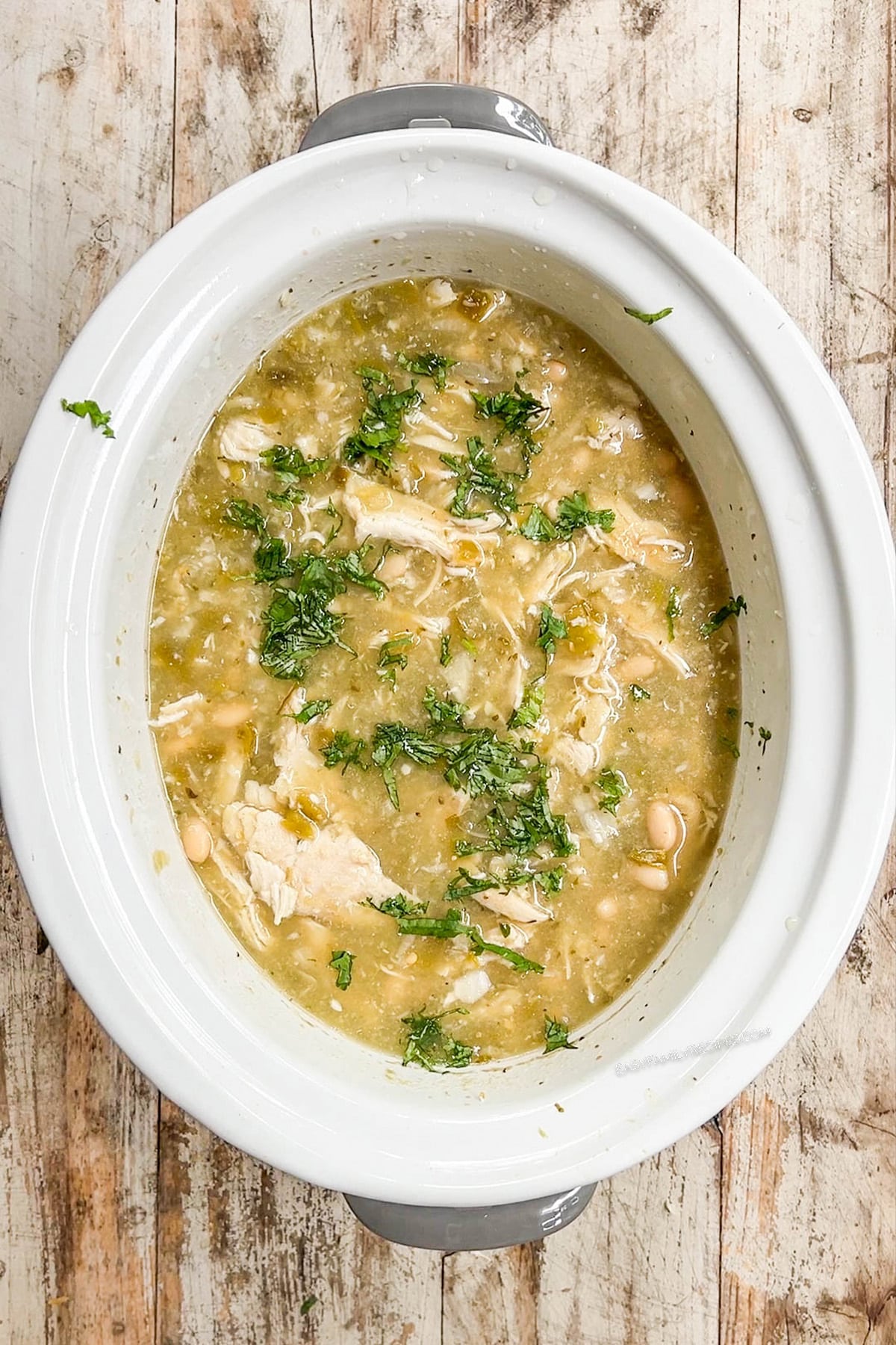 A top view of a white Crock Pot with the cooked chicken for Salsa Verde Soup. The chicken is shredded and the soup is ready to serve. Step 6 of the recipe.