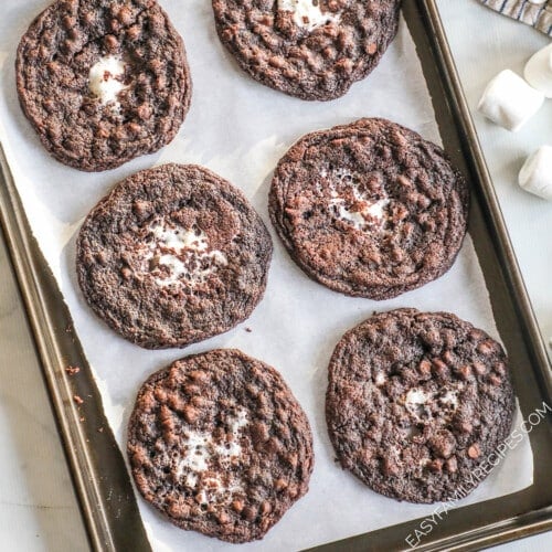 A top view of a dark silver baking sheet lined with parchment paper, and six chocolate marshmallow cookies.