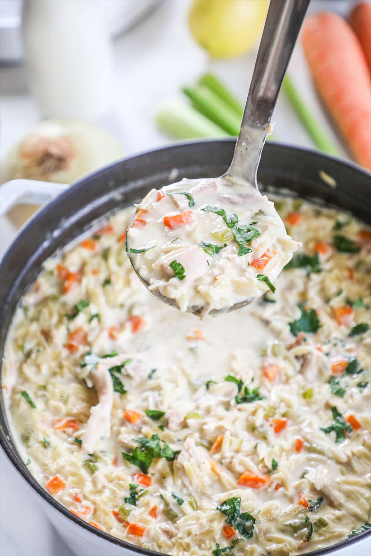 A ladle scoops out a portion of Creamy Lemon Chicken Orzo Soup from a Dutch oven. Veggies are in the background. 